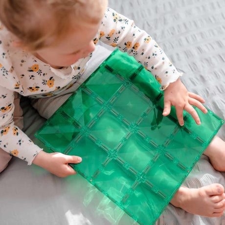 Toddler playing with a large green Connetix magnetic tile base plate while sitting on a bed.
