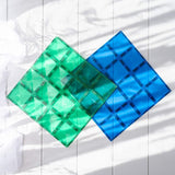 Translucent green and blue magnetic tile baseplates on a white wooden surface with soft leaf shadows.