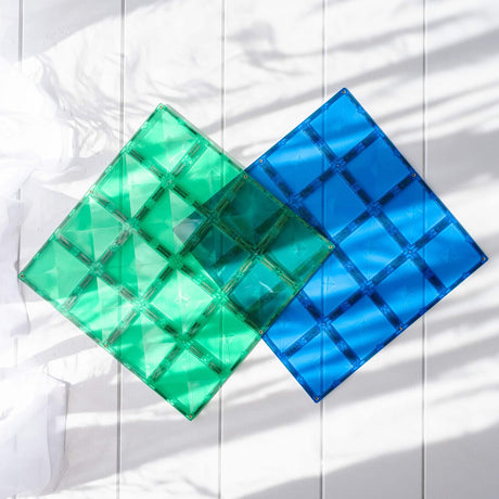 Translucent green and blue magnetic tile baseplates on a white wooden surface with soft leaf shadows.