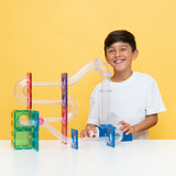 Smiling boy playing with a colorful magnetic tile ball run set against a bright yellow background.