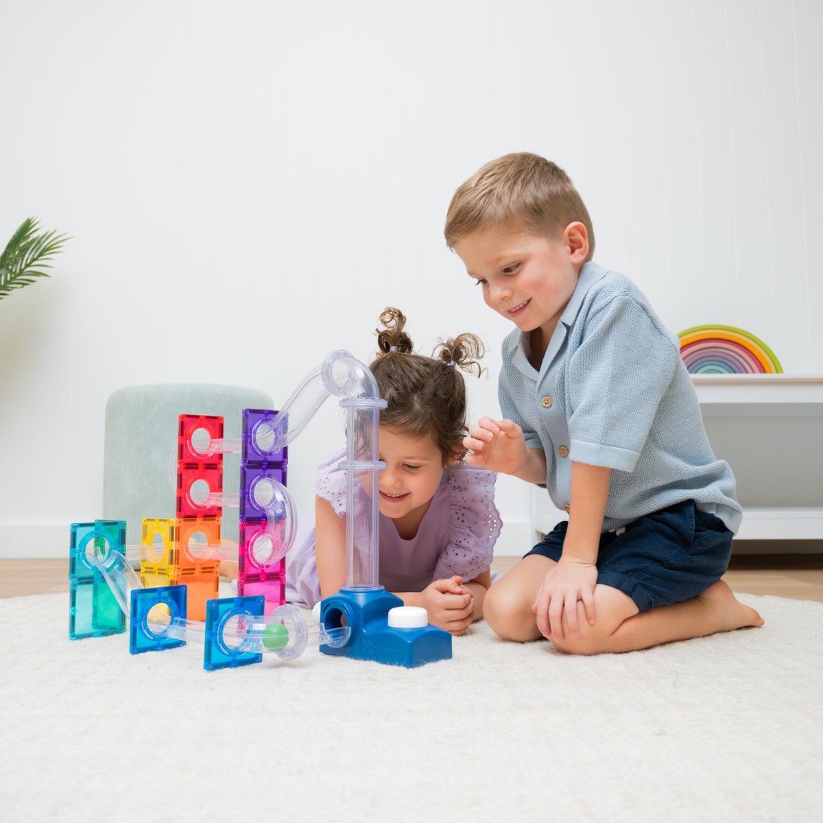 Two young children playing with a magnetic tile ball run structure on a white rug.