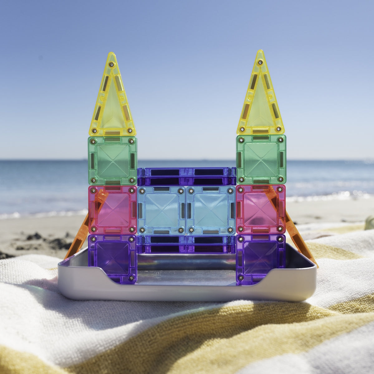 Colorful magnetic tile castle built on a tray on a beach towel with the ocean behind it.