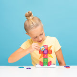 Young girl placing a bee-illustrated tile on a colorful magnetic tile flower against a blue backdrop.