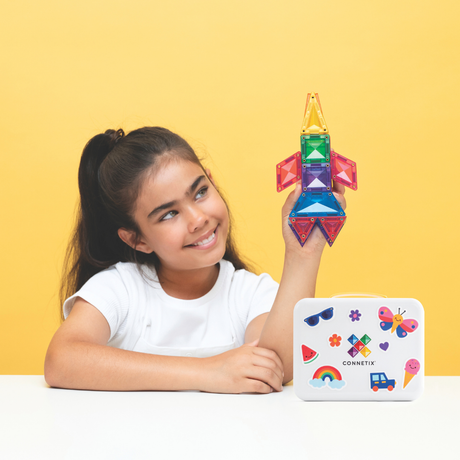 Young girl holding a colorful magnetic tile rocket next to a white carrying case on a yellow background.
