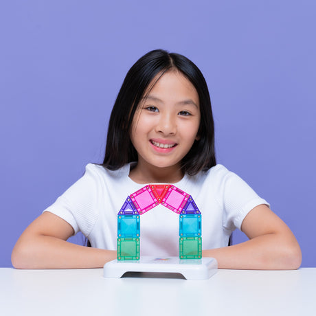 Smiling young girl sitting behind a colorful magnetic tile arch against a purple backdrop.