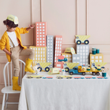 Child in a yellow cardigan standing on a chair next to a table set with construction-themed party supplies.