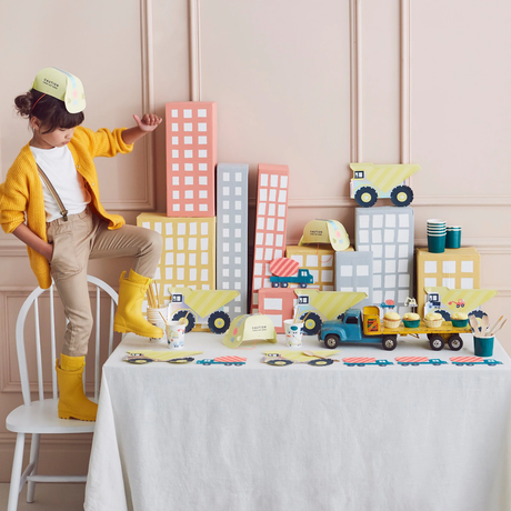 Child in a yellow cardigan standing on a chair next to a table set with construction-themed party supplies.