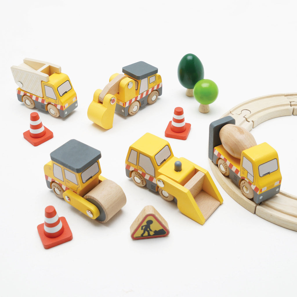 Wooden toy construction vehicles, including a dump truck, excavator, road roller, and cement mixer, are surrounded by traffic cones and a caution sign.