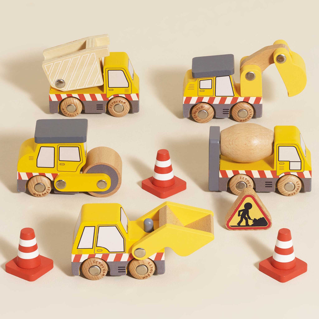 Wooden toy construction vehicles, including a dump truck, excavator, road roller, and cement mixer, are surrounded by traffic cones and a caution sign.