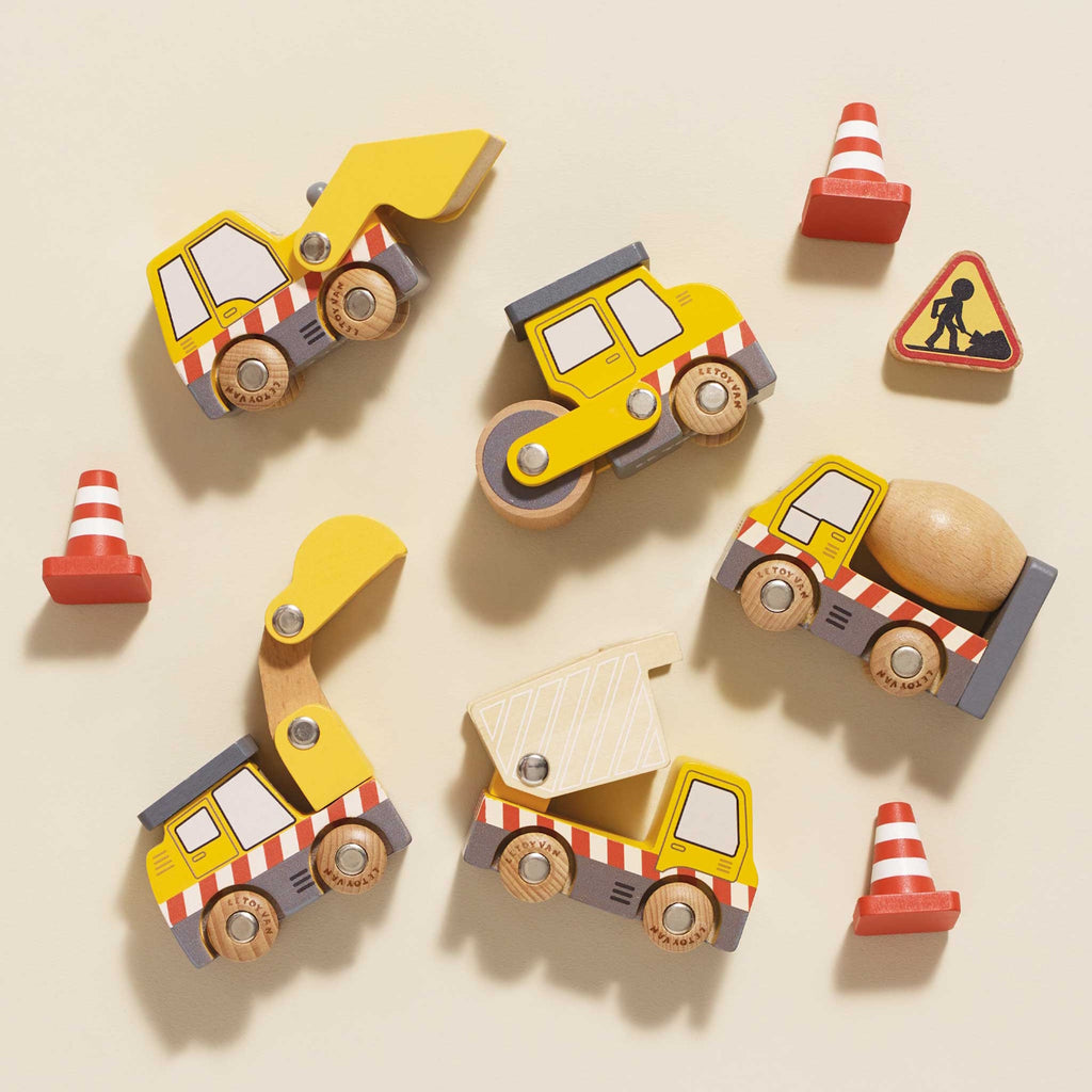 Wooden toy construction vehicles, including a dump truck, excavator, road roller, and cement mixer, are surrounded by traffic cones and a caution sign.