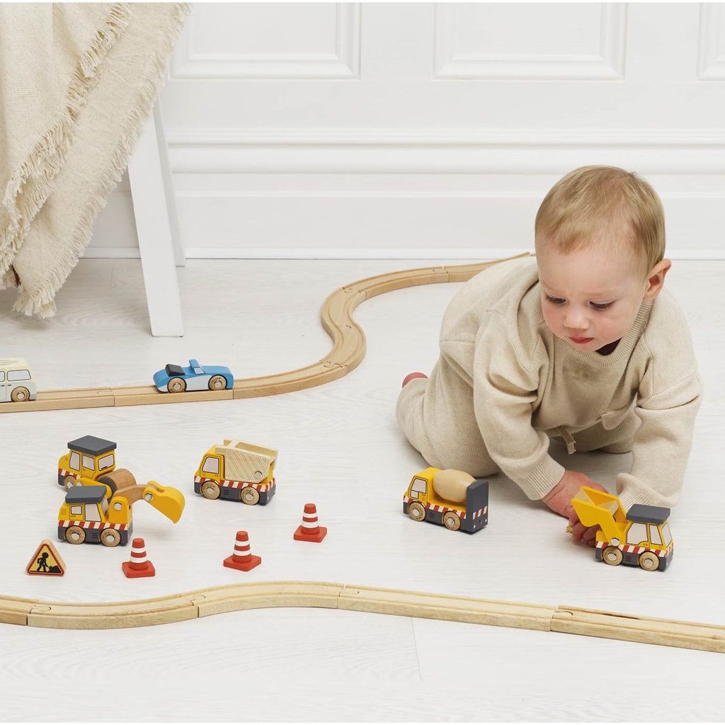 Wooden toy construction vehicles, including a dump truck, excavator, road roller, and cement mixer, are surrounded by traffic cones and a caution sign.