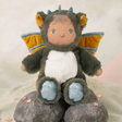 Cozy Day Dinkum Doll Flint wearing a fuzzy green dragon costume with wings, sitting on decorative rocks.