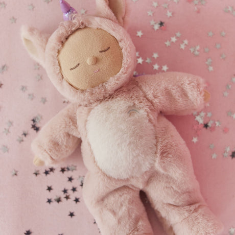 Olli Ella Cozy Dinkum Unicorn Sugarpie plush doll lying on a pink background with star confetti.