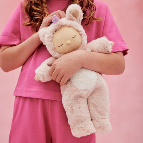 Child hugging an Olli Ella Cozy Dinkum Doll Unicorn Sugarpie plush toy against a pink background.