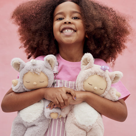 Smiling young girl hugging two plush Olli Ella Cozy Dinkum Dolls against a solid pink background.