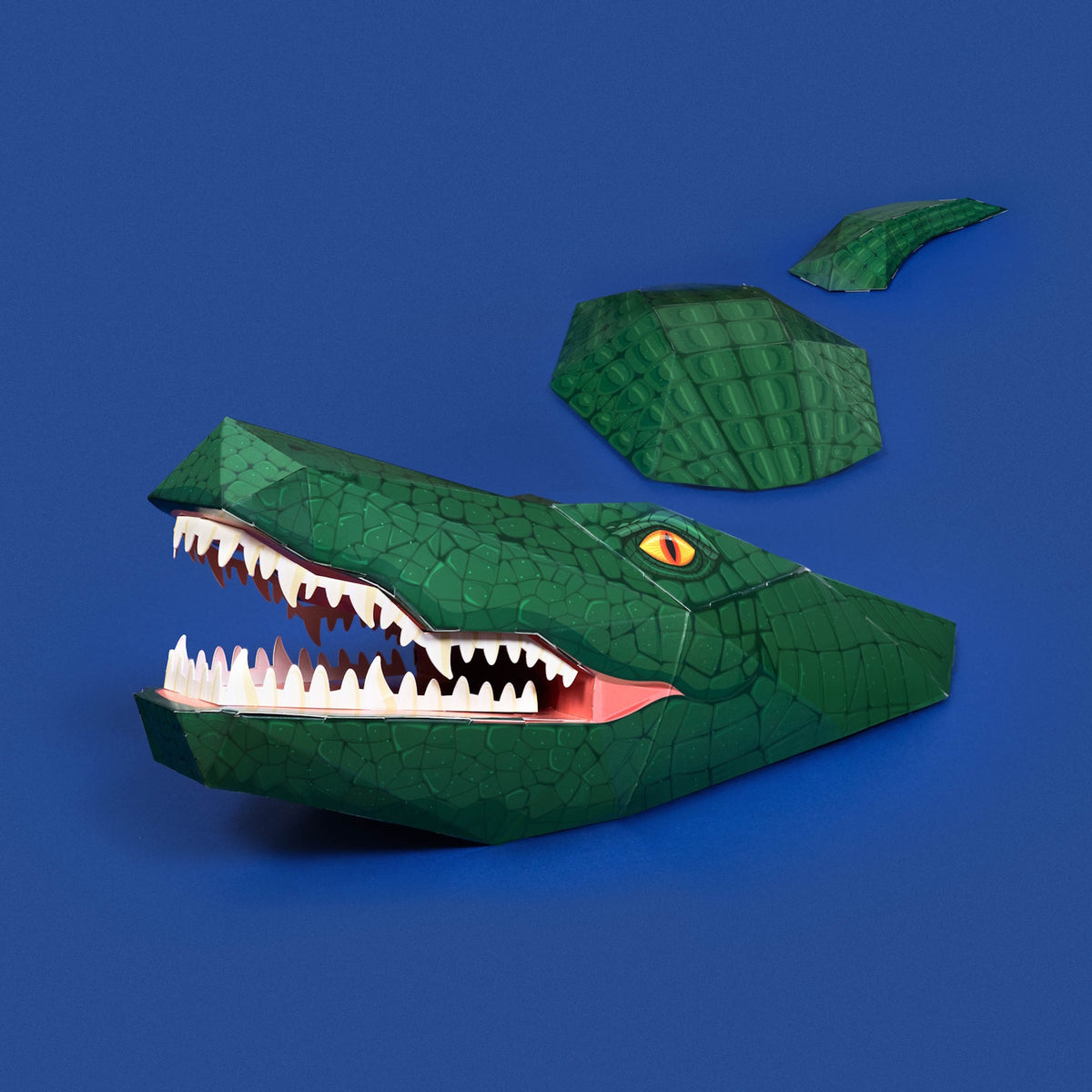 3D papercraft green crocodile head, back, and tail pieces arranged on a solid blue background.