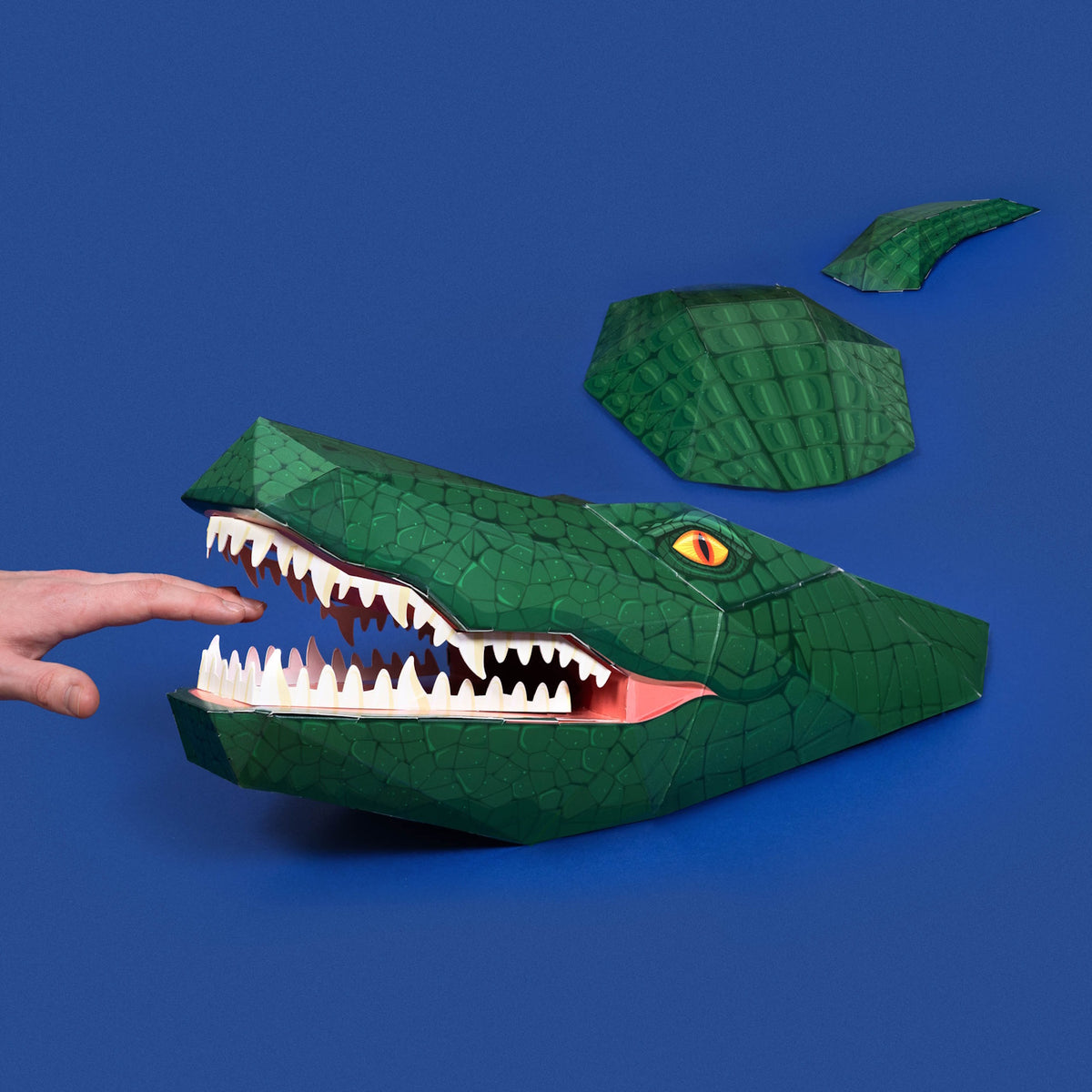 A green papercraft crocodile with its open mouth near a finger, against a solid blue background.