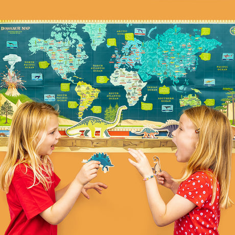 Two young girls excitedly playing with dinosaur cutouts in front of a large, colorful dinosaur world map.
