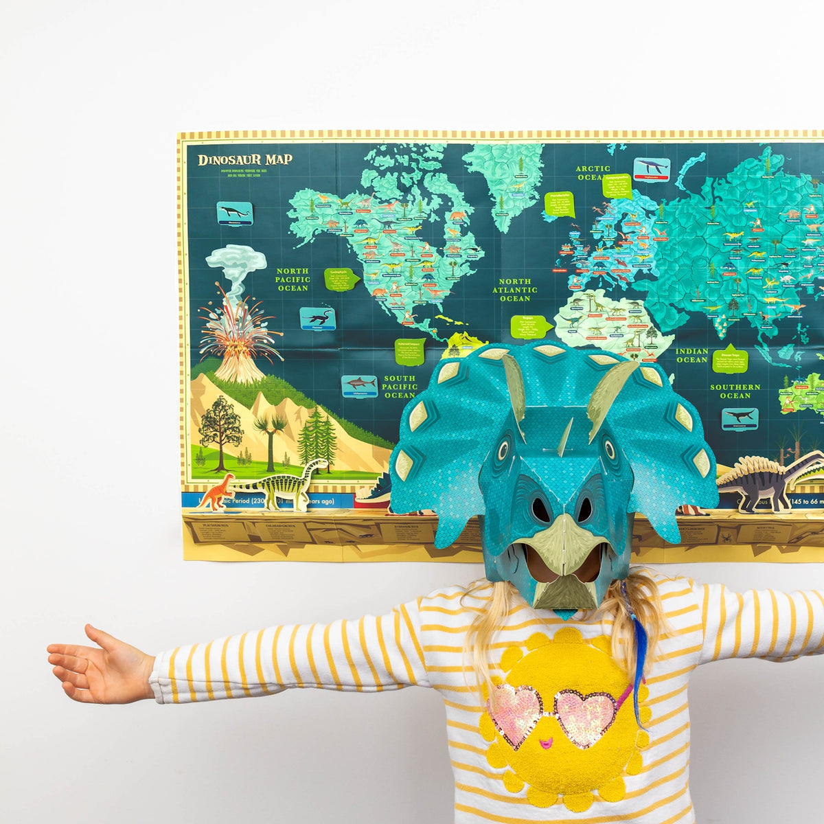 Child wearing a turquoise dinosaur mask with arms outstretched in front of a dinosaur world map poster.