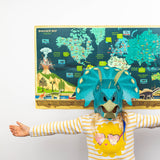 Child wearing a turquoise dinosaur mask with arms outstretched in front of a dinosaur world map poster.