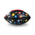 A black football features colorful illustrations of planets, rockets, and stars, with vibrant red stitching, conveying a playful space-themed design.