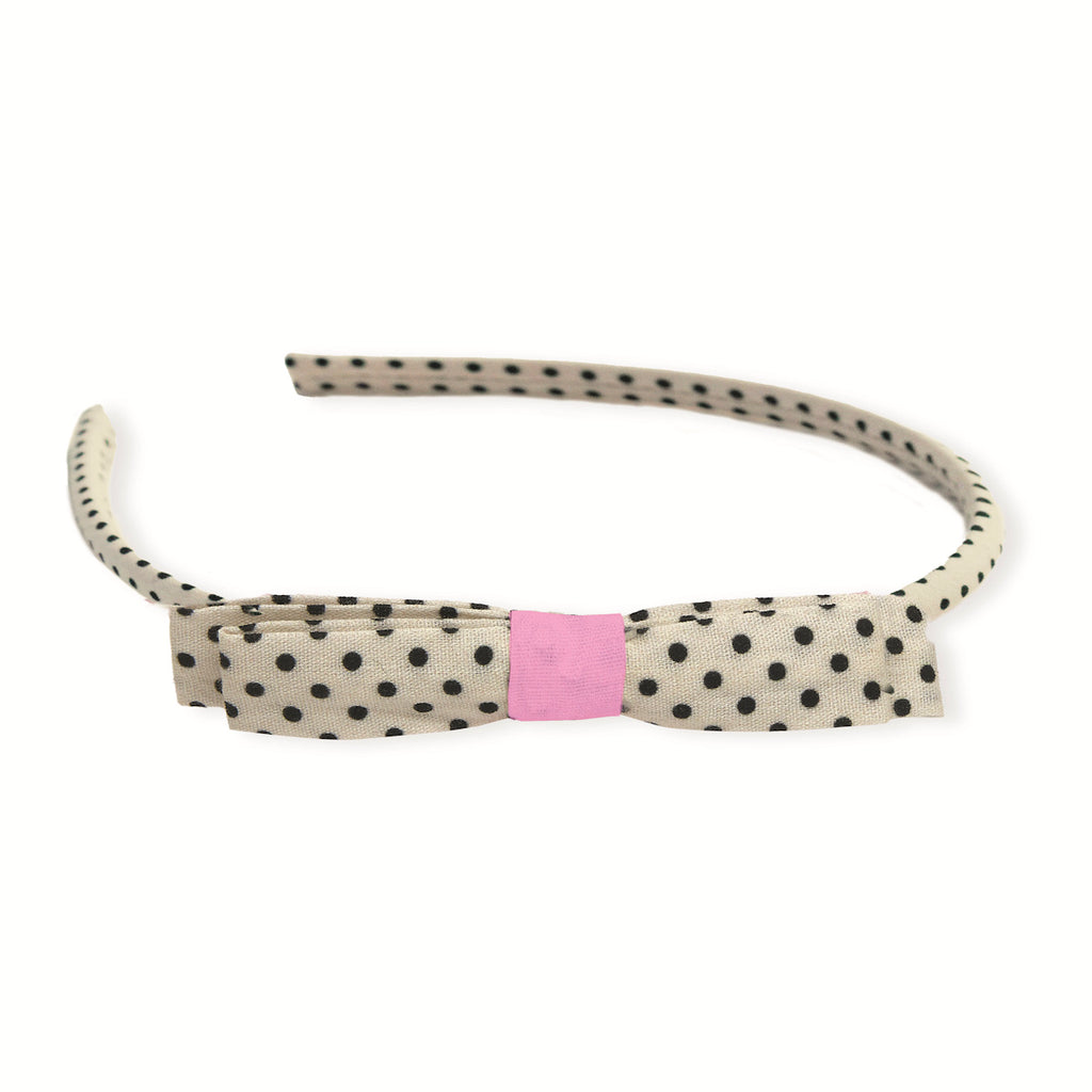 Polka-dotted headband in beige with black dots, featuring a cute bow and pink fabric accent in the center. Playful and stylish accessory.