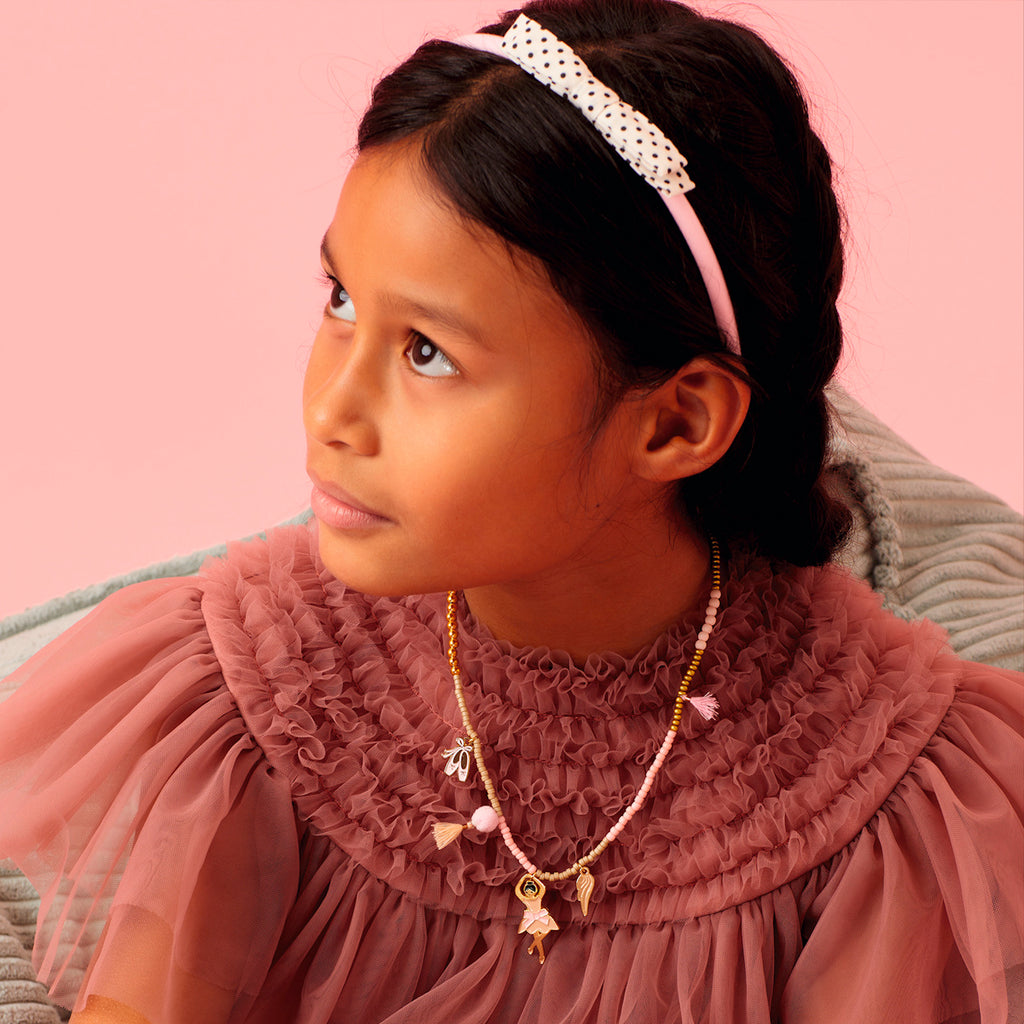 Young girl in a textured pink dress and beaded necklace with charms and dotted headband, looking down thoughtfully. Soft pink background enhances a serene mood.