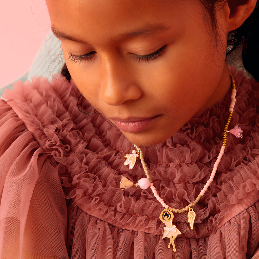 Young girl in a textured pink dress and beaded necklace with charms, looking down thoughtfully. Soft pink background enhances a serene mood.