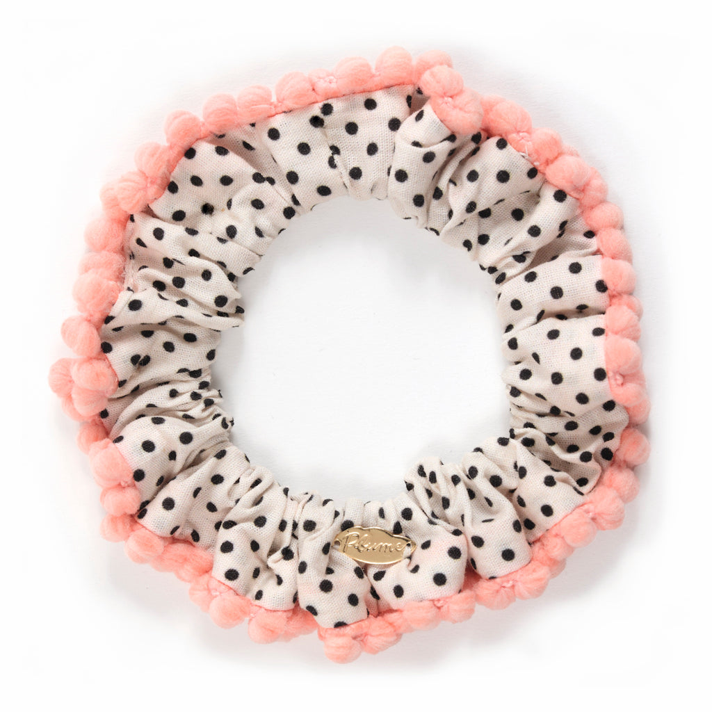 A fabric scrunchie with black polka dots on a white background, adorned with a pink pom-pom trim. A small gold tag is attached, adding a playful, chic touch.