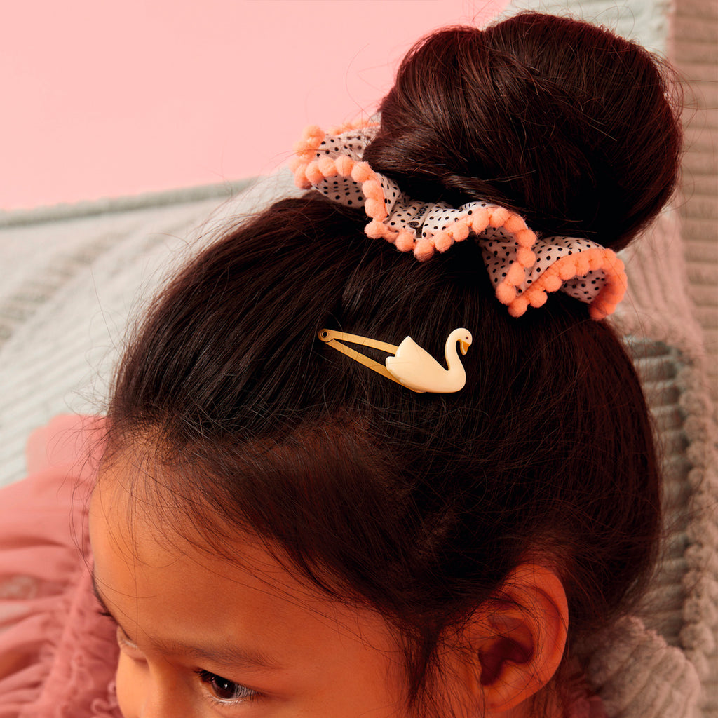 Young girl with dark hair in a bun, adorned with a pink scrunchie and a swan-shaped clip. The background is soft pink, creating a playful tone.