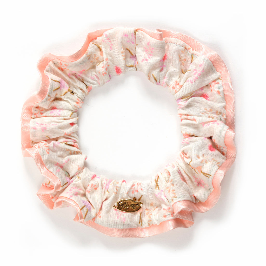 Floral scrunchie with pastel pink and white fabric, featuring a delicate pink blossoms pattern and a small gold leaf charm. Soft, feminine design.