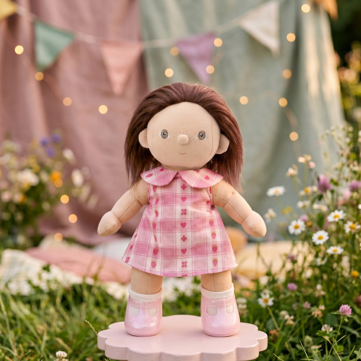 Dinkum doll wearing a pink plaid rosette dress and matching pink shoes in an outdoor garden setting.