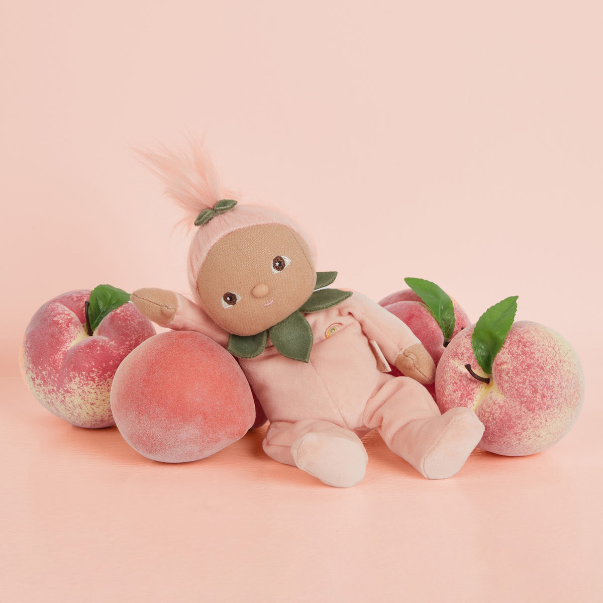 Olli Ella Dinky Dinkums Peggy Peach plush doll sitting among fresh peaches.