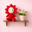 Red flower Dinky Dinkum doll sitting next to a potted plant with colorful daisies on a wooden shelf.