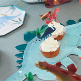 Cupcakes with white frosting and dinosaur toppers displayed on a dinosaur-shaped serving platter alongside party plates.