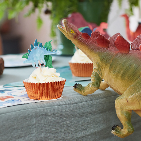 Large toy dinosaur overlooking a cupcake with a blue dinosaur topper in an orange foil baking cup on a table.