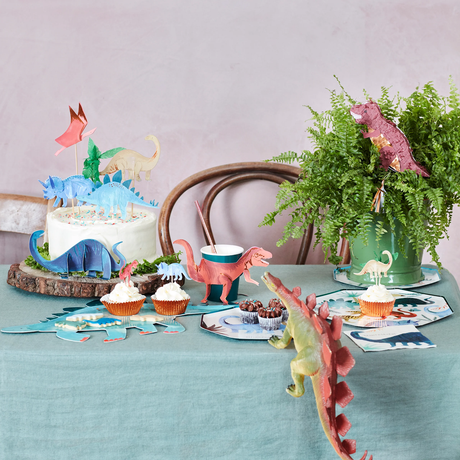 Table set with dinosaur-themed party decorations, including a frosted cake, cupcakes with dinosaur toppers, and toy dinosaurs.