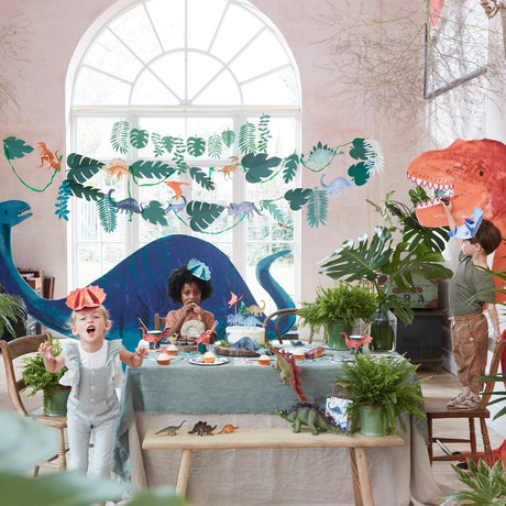 Children at a dinosaur-themed party room decorated with the dinosaur garland, large dinosaur cutouts, and party table settings.