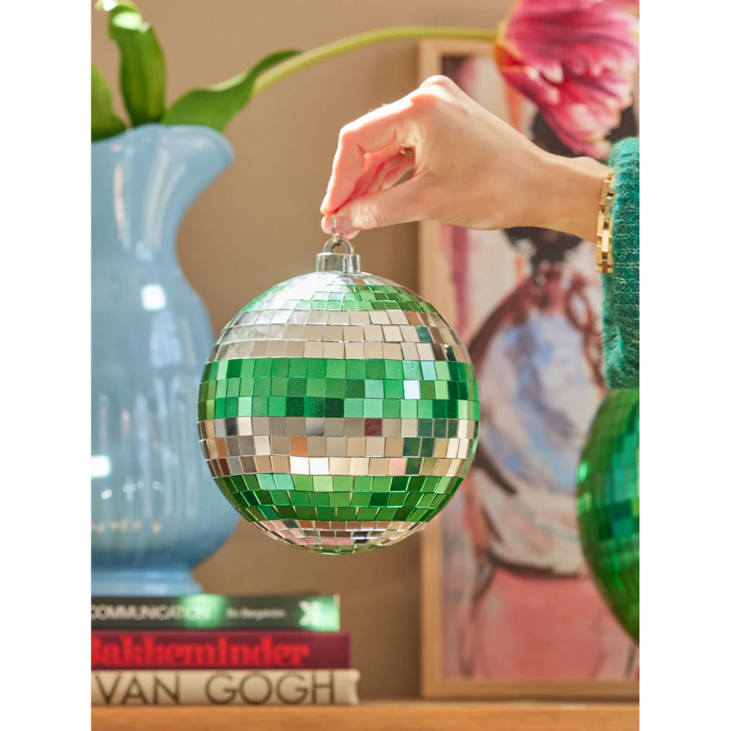 A hand holds a green and silver disco ball ornament. Nearby, a blue vase houses green leaves, and art books are stacked on a wooden surface. The mood is festive.