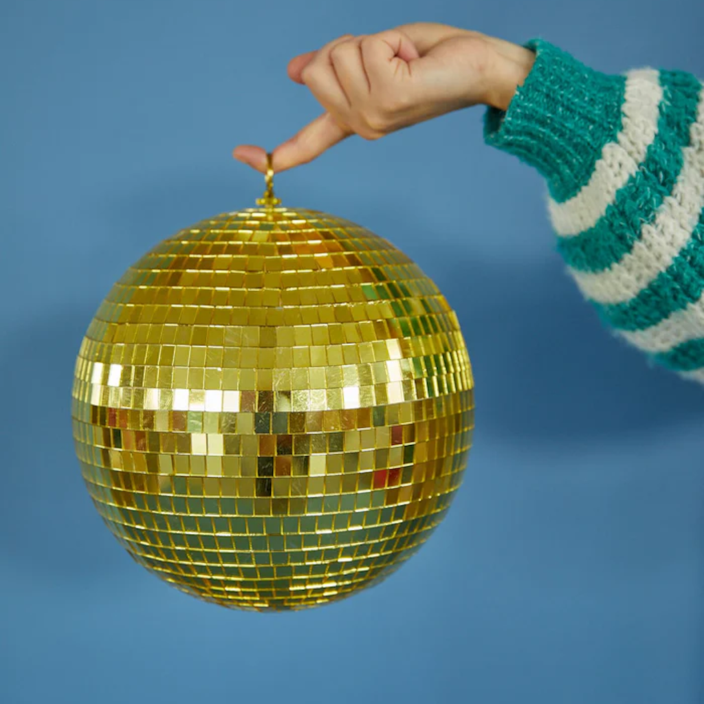 A hand in a green and white striped sweater holds a golden disco ball by its top loop against a solid blue background, conveying a playful, retro vibe.