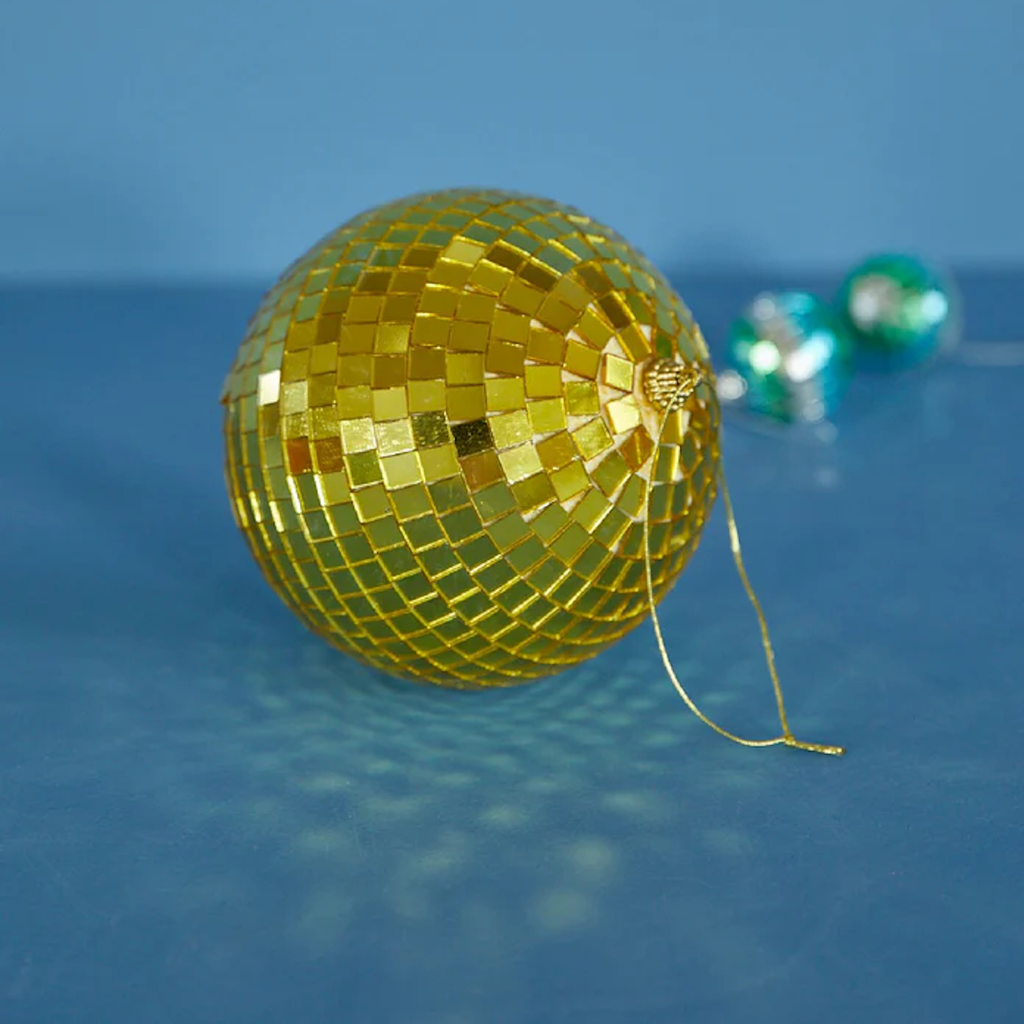 A shiny golden disco ball ornament with a reflective mosaic pattern rests on a blue surface. Two blurred teal ornaments are seen in the background.