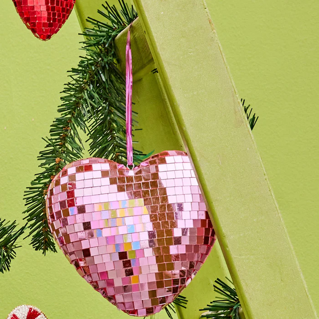 A pink disco ball heart ornament hangs from a green ladder with pine garland. The background is a light green, creating a festive and vibrant atmosphere.