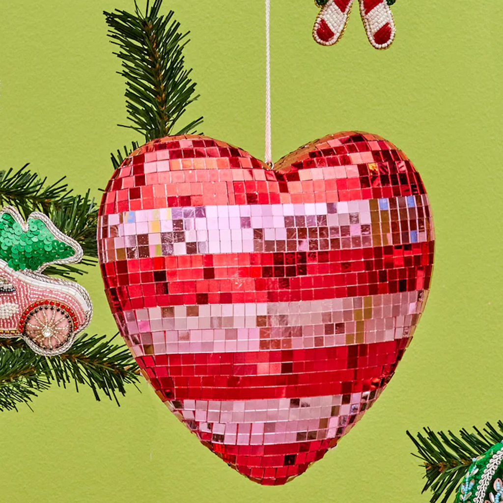 Sparkling heart-shaped ornament in red and pink hangs against a light green wall. Surrounded by festive decorations and evergreen branches.