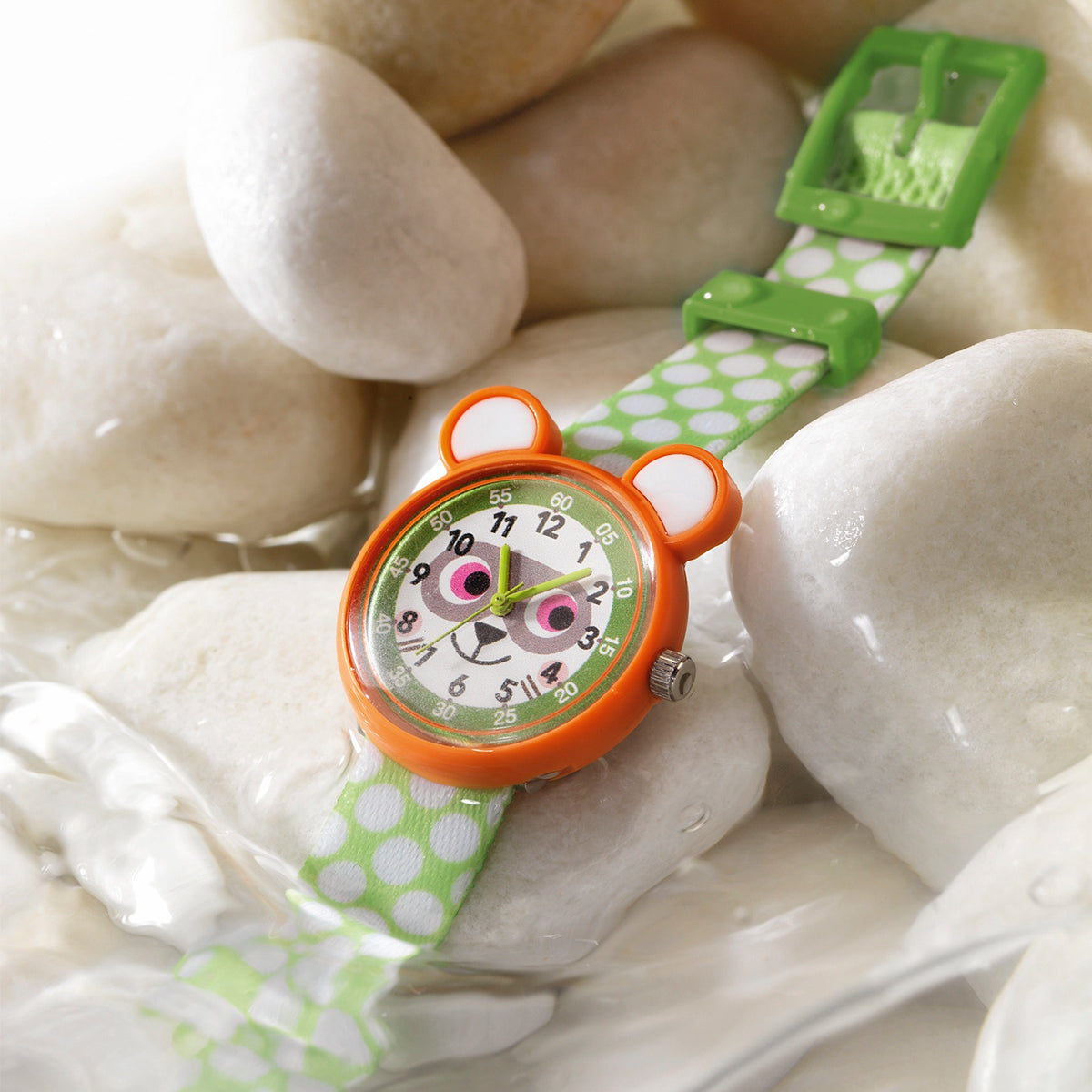 Raccoon-themed children's watch with a green polka-dot strap resting on smooth white stones in water.