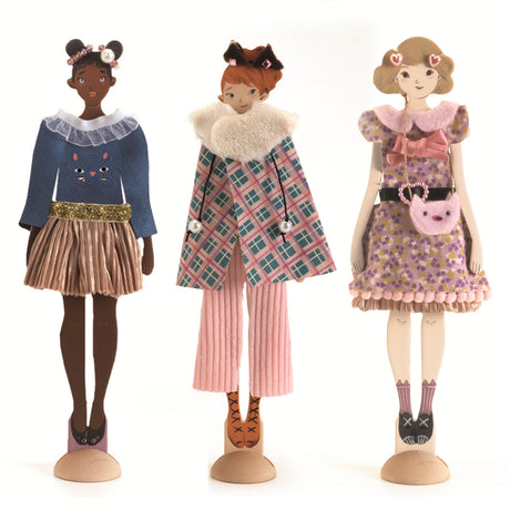 Three stylish dolls stand on wooden bases, each showcasing unique outfits with vibrant colors and playful accessories.