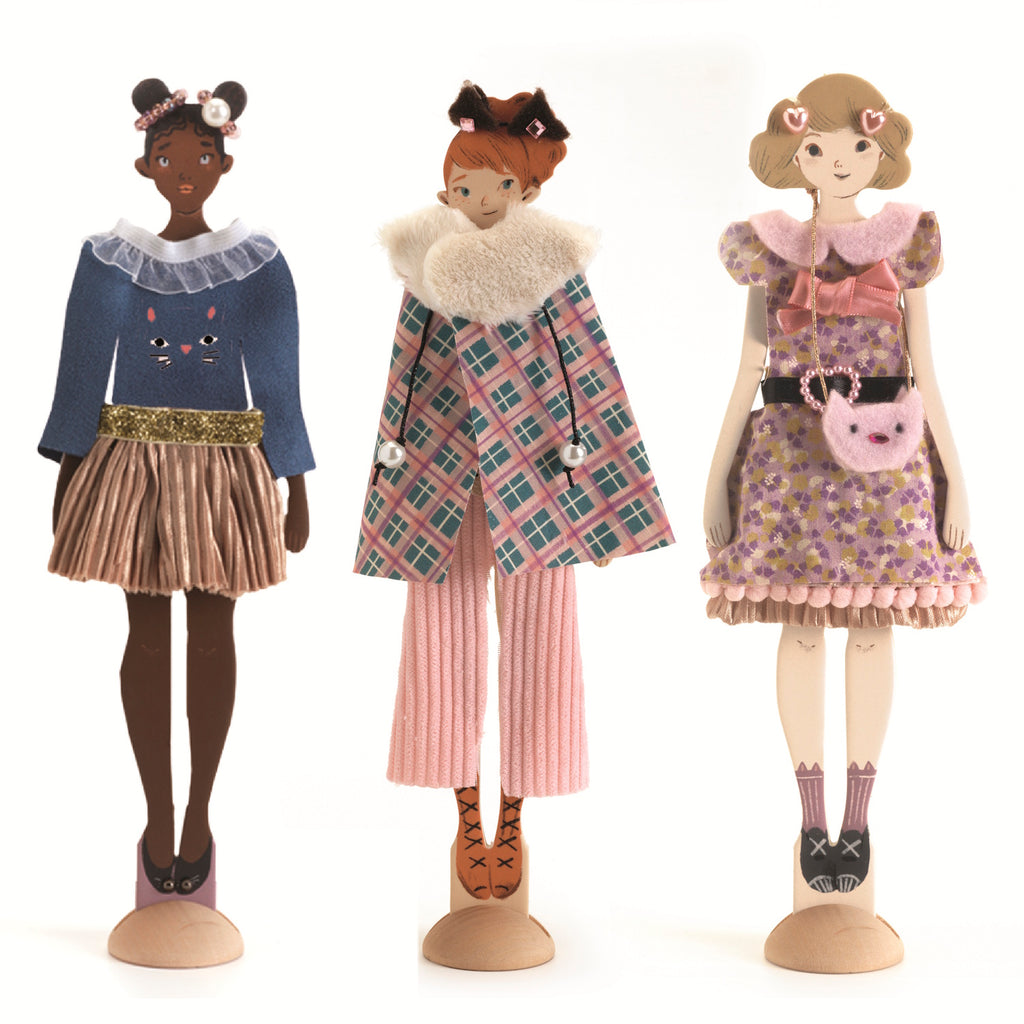 Three stylish dolls stand on wooden bases, each showcasing unique outfits with vibrant colors and playful accessories.
