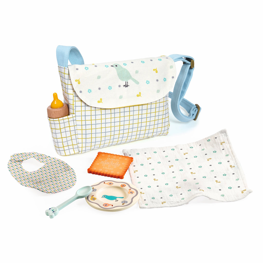 A diaper bag with a blue strap features a bird design, checkered pattern, and contains a toy bottle. Arranged beside it are a bib, cookie toy, spoon, plate, and patterned cloth, conveying a cheerful and playful tone.