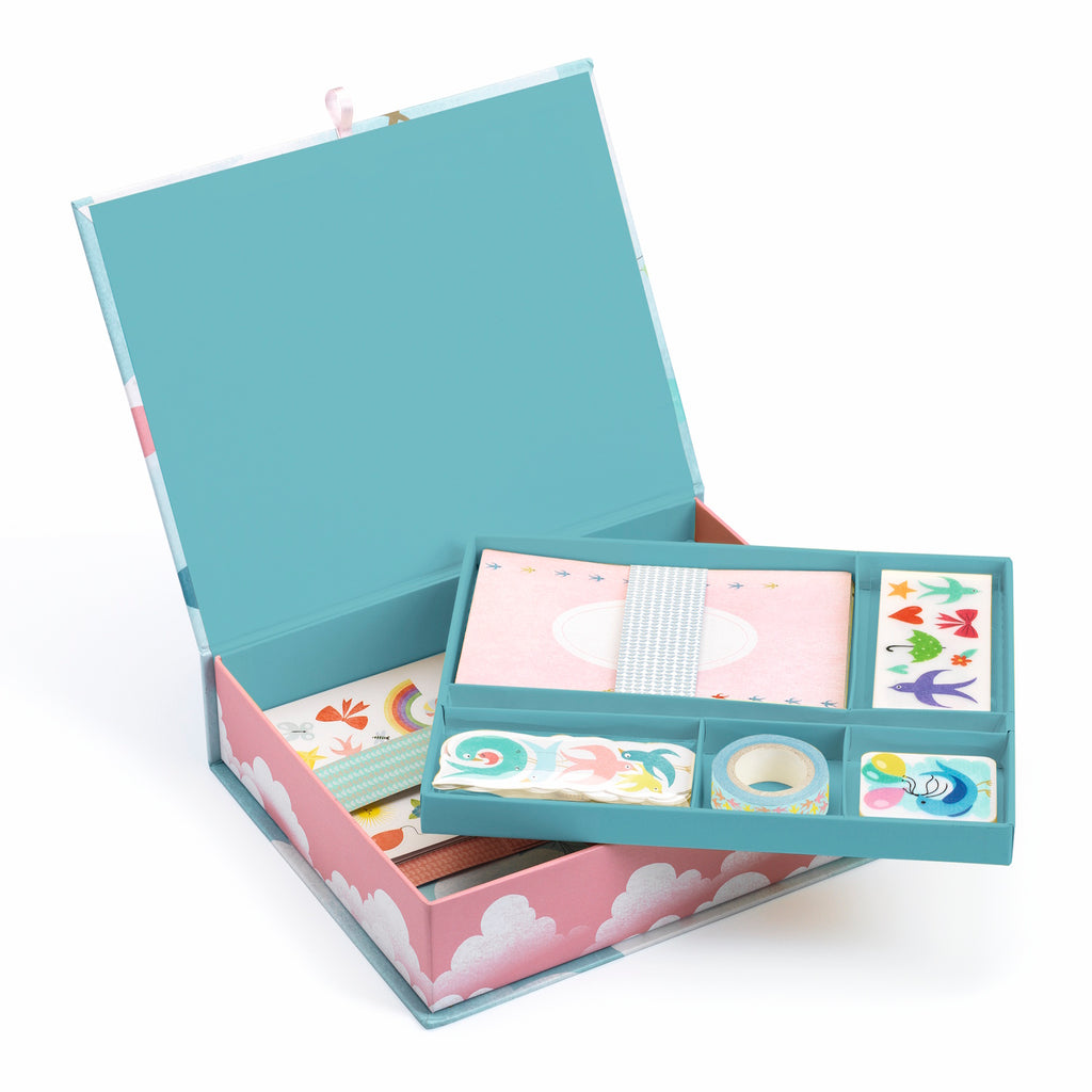 An open stationery box with a teal lid and pink cloud-patterned exterior reveals colorful items inside, including paper, stickers, and decorative tape.