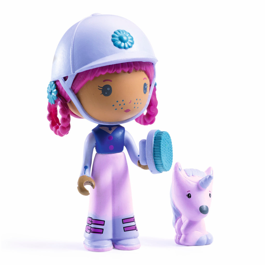 A doll with pink hair and a white helmet, holding a blue brush, stands beside a small purple unicorn. The doll wears a blue and pink outfit.
