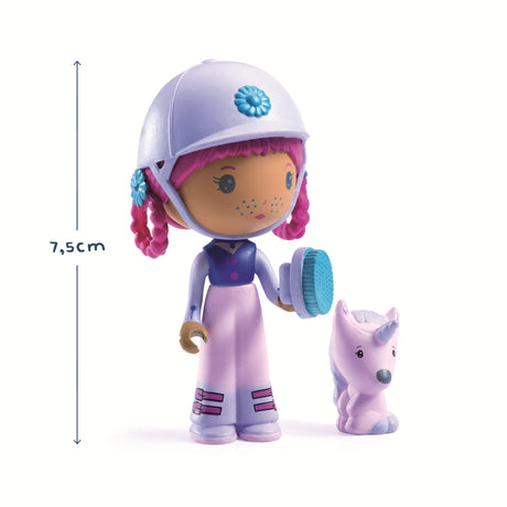 A doll with pink hair and a white helmet, holding a blue brush, stands beside a small purple unicorn. The doll wears a blue and pink outfit.